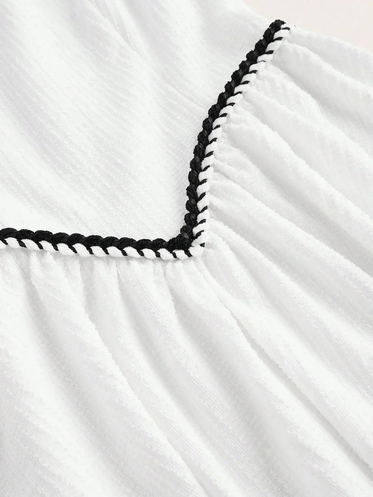 This charming white dress features delicate textured fabric with a playful black braided trim along the neckline, adding a.