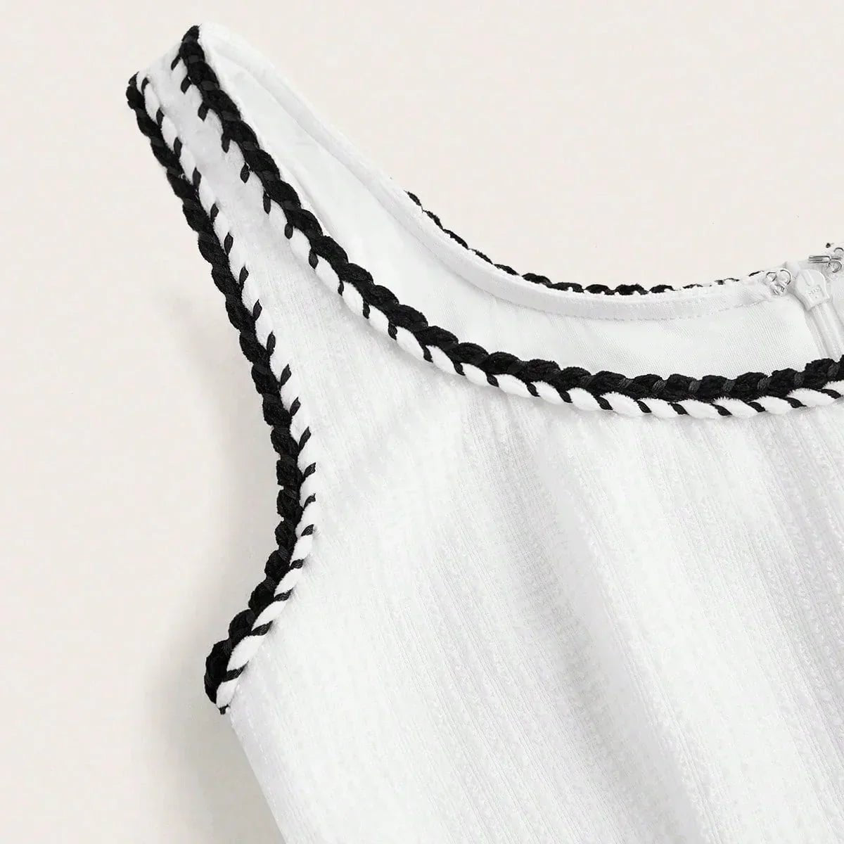 This stunning sleeveless dress features a crisp white fabric accentuated by chic black braided trim along the neckline,.