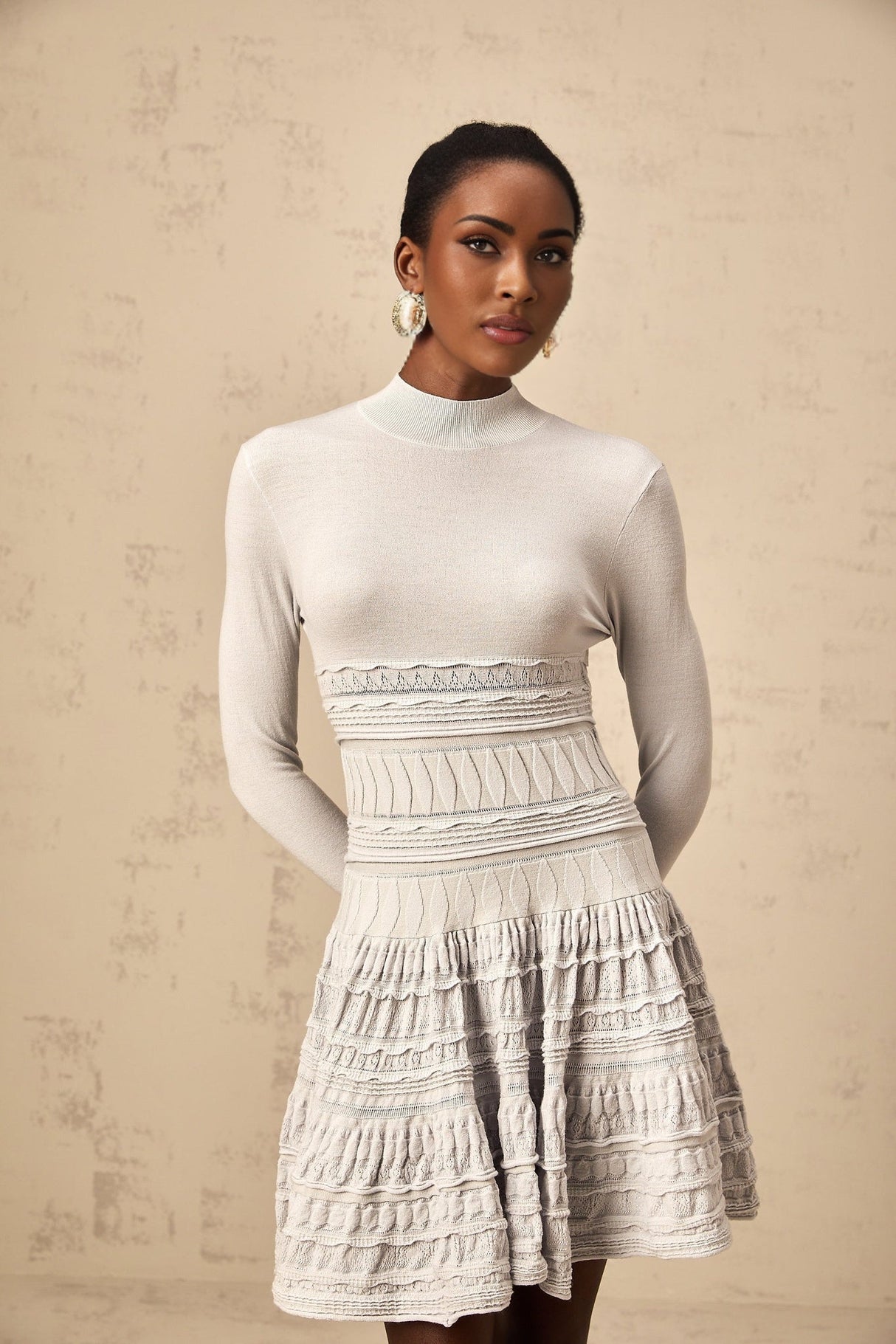 Woman wearing a short grey dress with ruffled details and ribbed texture standing against plain background
