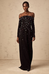 A black crystalembellished offshoulder maxi dress with a long flowing skirt worn by a model against a plain background