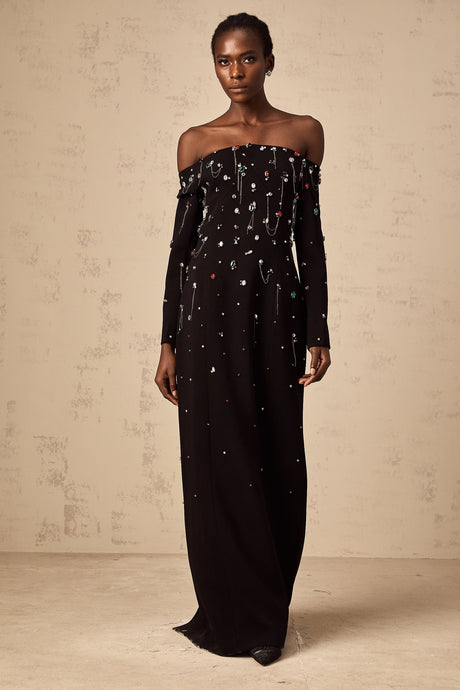 A black crystalembellished offshoulder maxi dress with a long flowing skirt worn by a model against a plain background