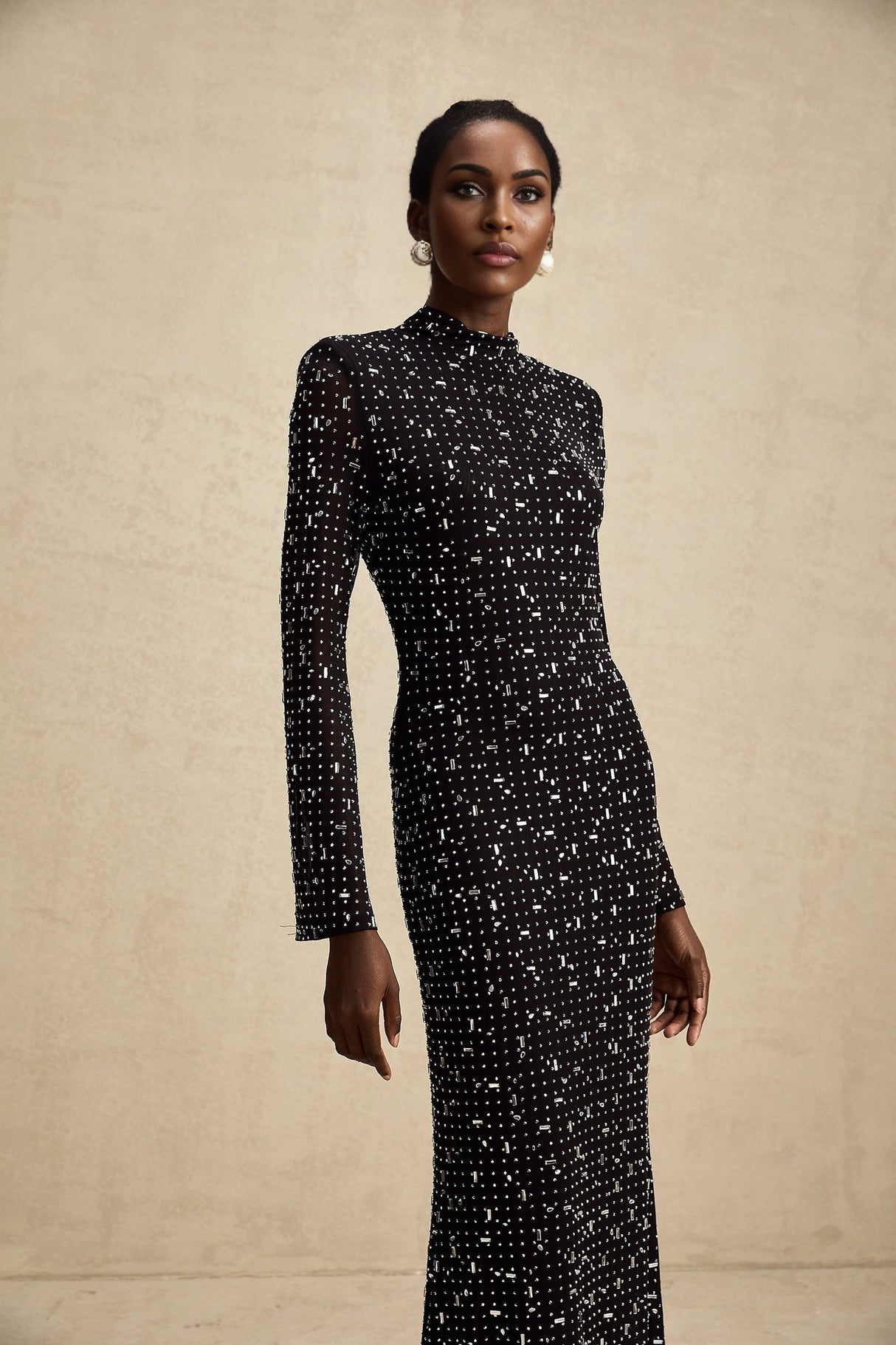 A black floor length maxi dress with sheer elements and decorative embellishments worn by a model