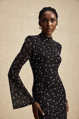 A black floor length semi sheer maxi dress with embellishments worn by a model against a plain background