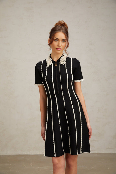 A black knit mini dress with vertical white stripes and ruffle trim details worn by a model standing against a plain background