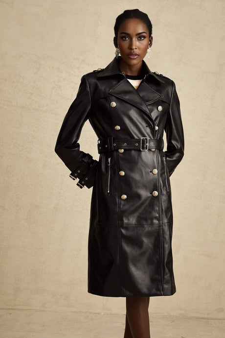 A black leather coat with doublebreasted front closure and a belt cinching the waist worn by a model against a plain background