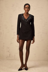 A black mini dress with a plunging V neckline featuring crystal embellishments worn by a model against a plain background