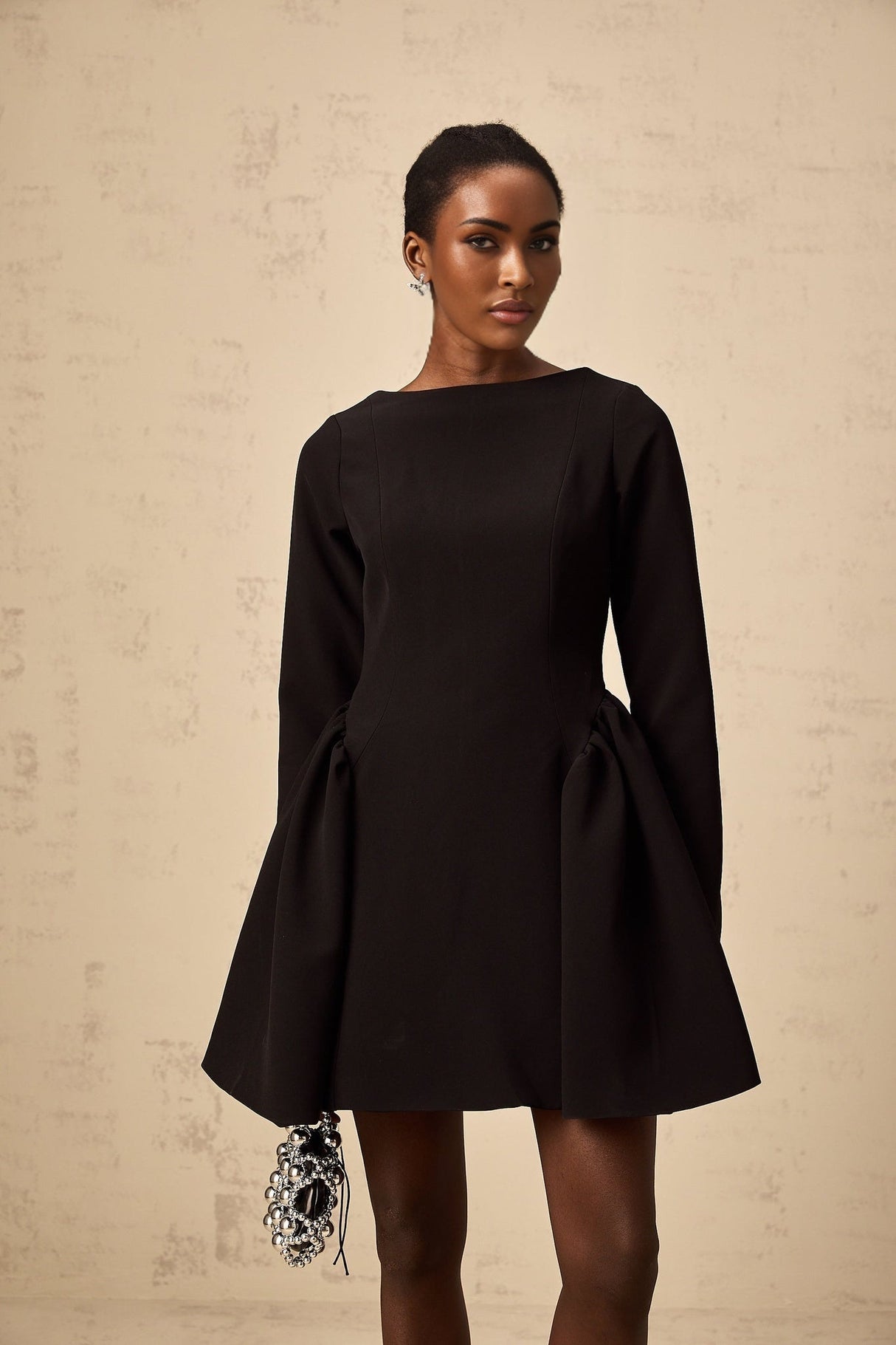 A black mini dress with long sleeves and a flared skirt worn by a model standing against a plain background