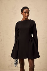 A black mini dress with long sleeves and a flared skirt worn by a model standing against a plain background