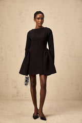 A black mini dress with long sleeves and a flared skirt worn by a model standing against a plain background
