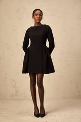 A black mini dress with long sleeves and a flared skirt worn by a model standing against a plain background