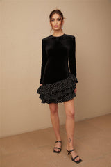 A black velvet mini dress with white polka dots featuring ruffled panels and a short hemline The dress has a vintage inspired look with a fitted silhouette and contrasting textures