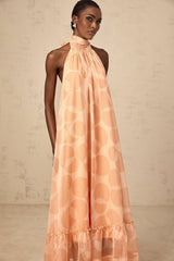 A coralpink halterneck maxi dress with a pattern print worn by a model named Clemence featuring a long flowing skirt and sleeveless design