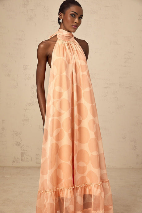 A coralpink halterneck maxi dress with a pattern print worn by a model named Clemence featuring a long flowing skirt and sleeveless design
