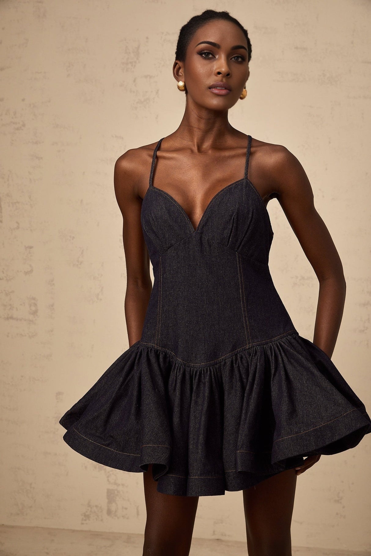 A dark blue denim mini dress with flounce details worn by a model against a plain background