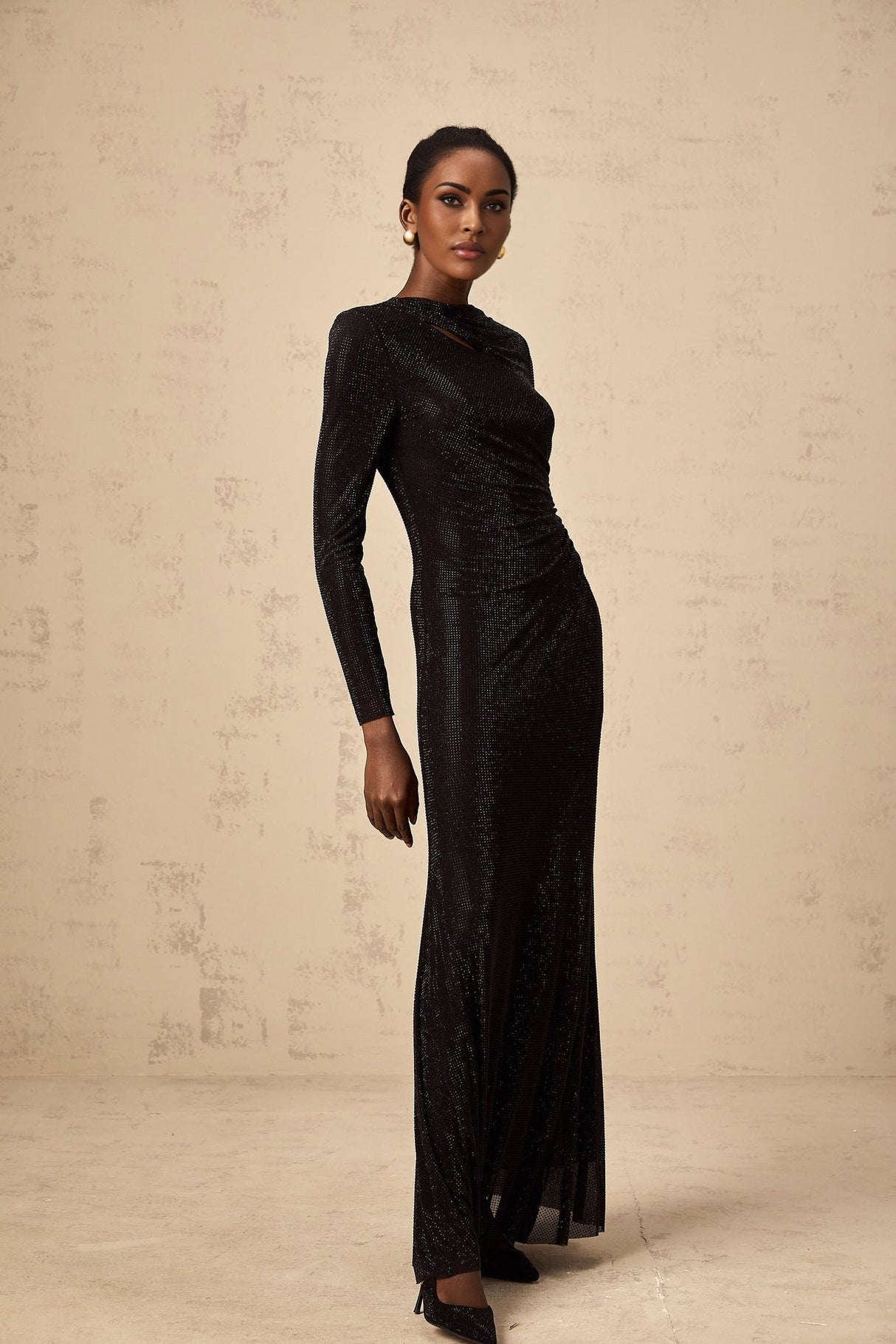 A long black maxi dress with ruched mesh fabric and rhinestone embellishments covering the bodice and sleeves