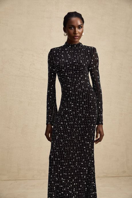 A long black semi sheer maxi dress with embellishments worn by a model standing against a plain background