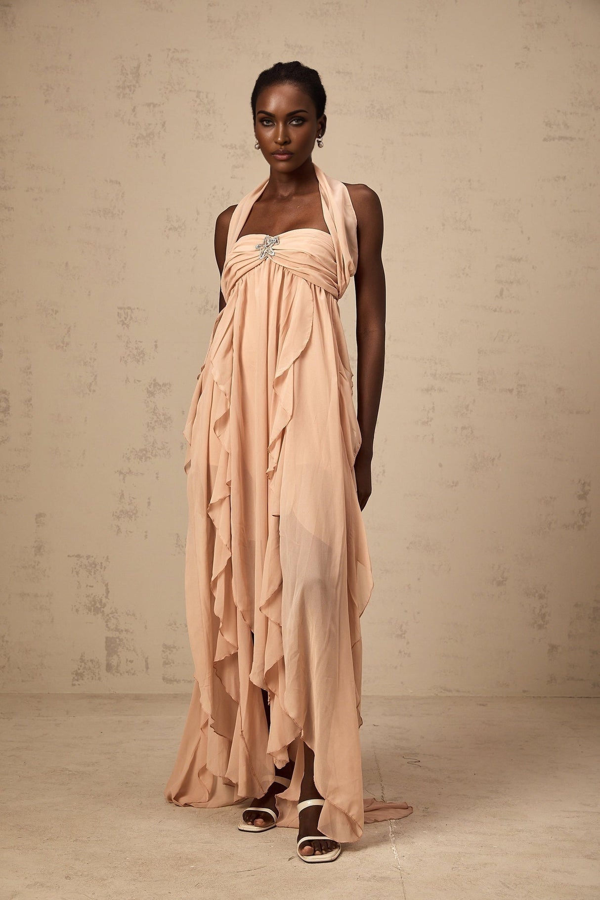 A long pink maxi dress with a halter neck and ruffled chiffon details worn by a model standing against a plain background