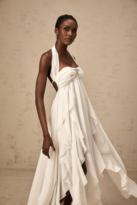A long white chiffon maxi dress with ruffle details and a halter neckline worn by a model against a plain background