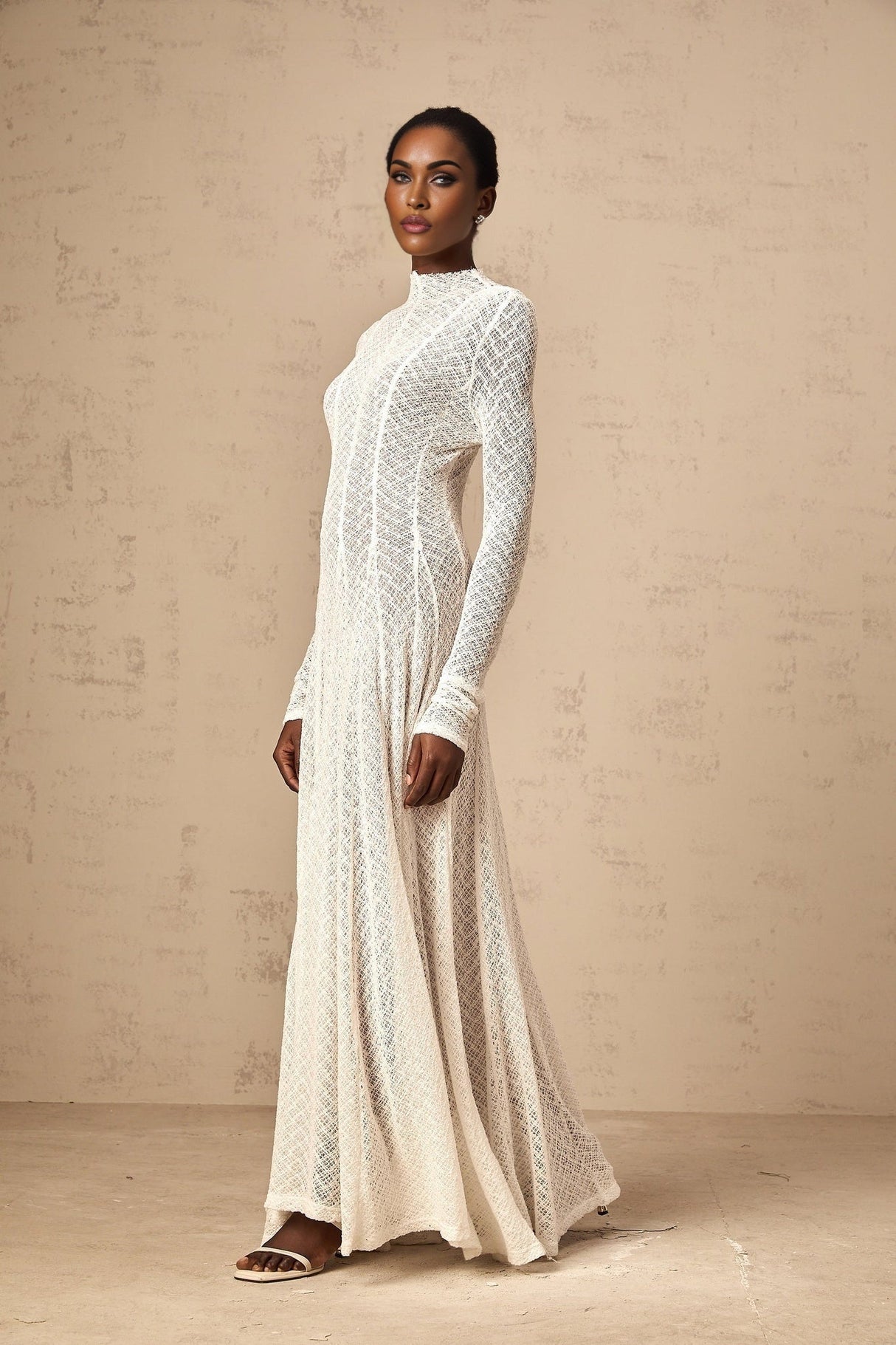 A long white lace maxi dress with a wavy pattern design worn by a model standing in a neutral setting