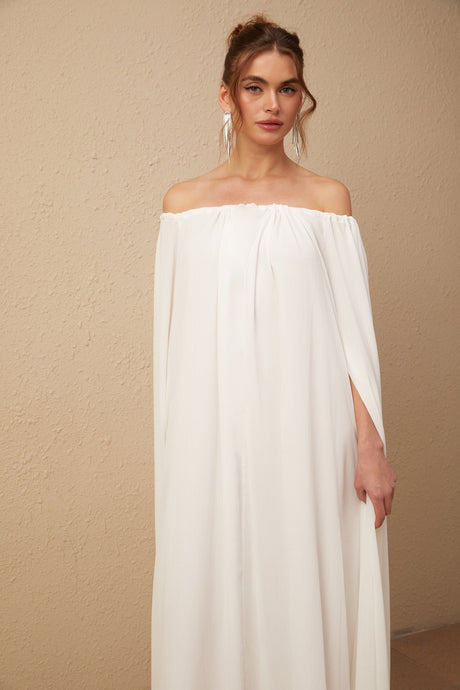 A long white maxi dress with a flowy tulle skirt and a capelike design draping over the shoulders creating an elegant silhouette