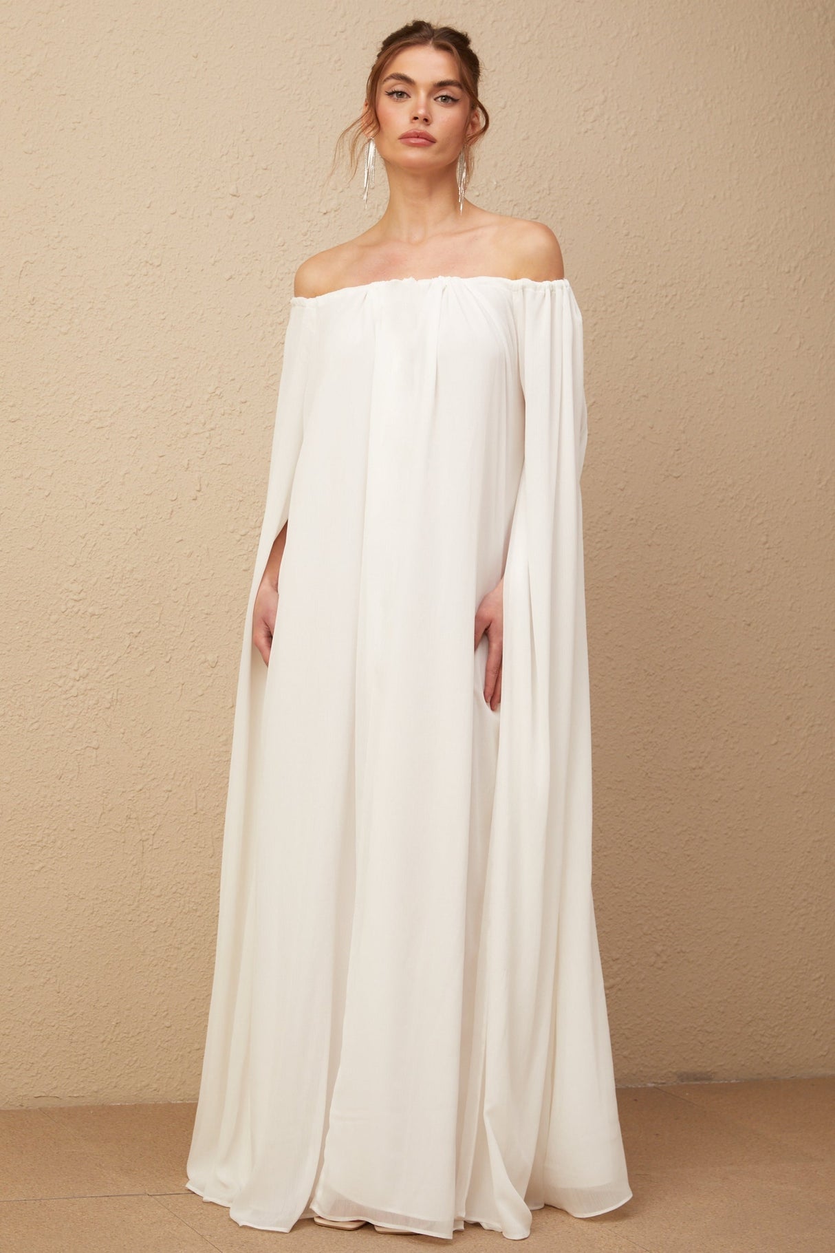 A long white tulle maxi dress with a draped cape design worn by a model standing against a plain background