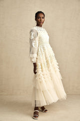 A long white tulle maxi dress with multiple layers featuring delicate floral embroidery in vibrant colors layered over a fitted underdress with a high neckline and long sleeves