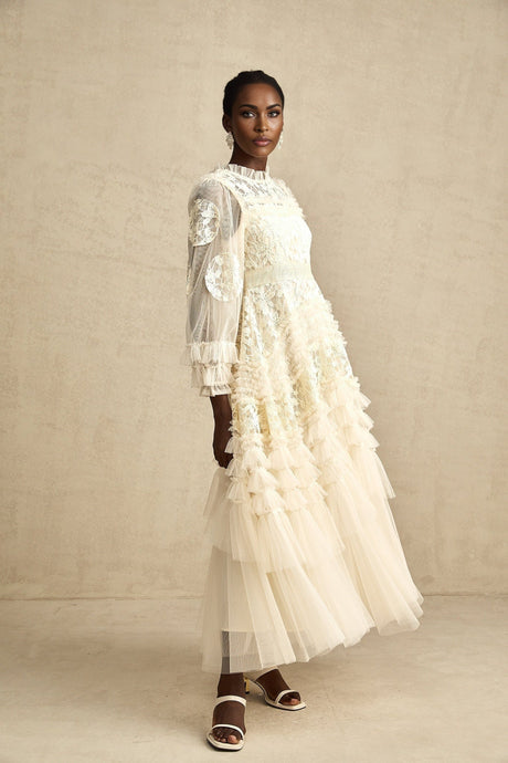 A long white tulle maxi dress with multiple layers featuring delicate floral embroidery in vibrant colors layered over a fitted underdress with a high neckline and long sleeves