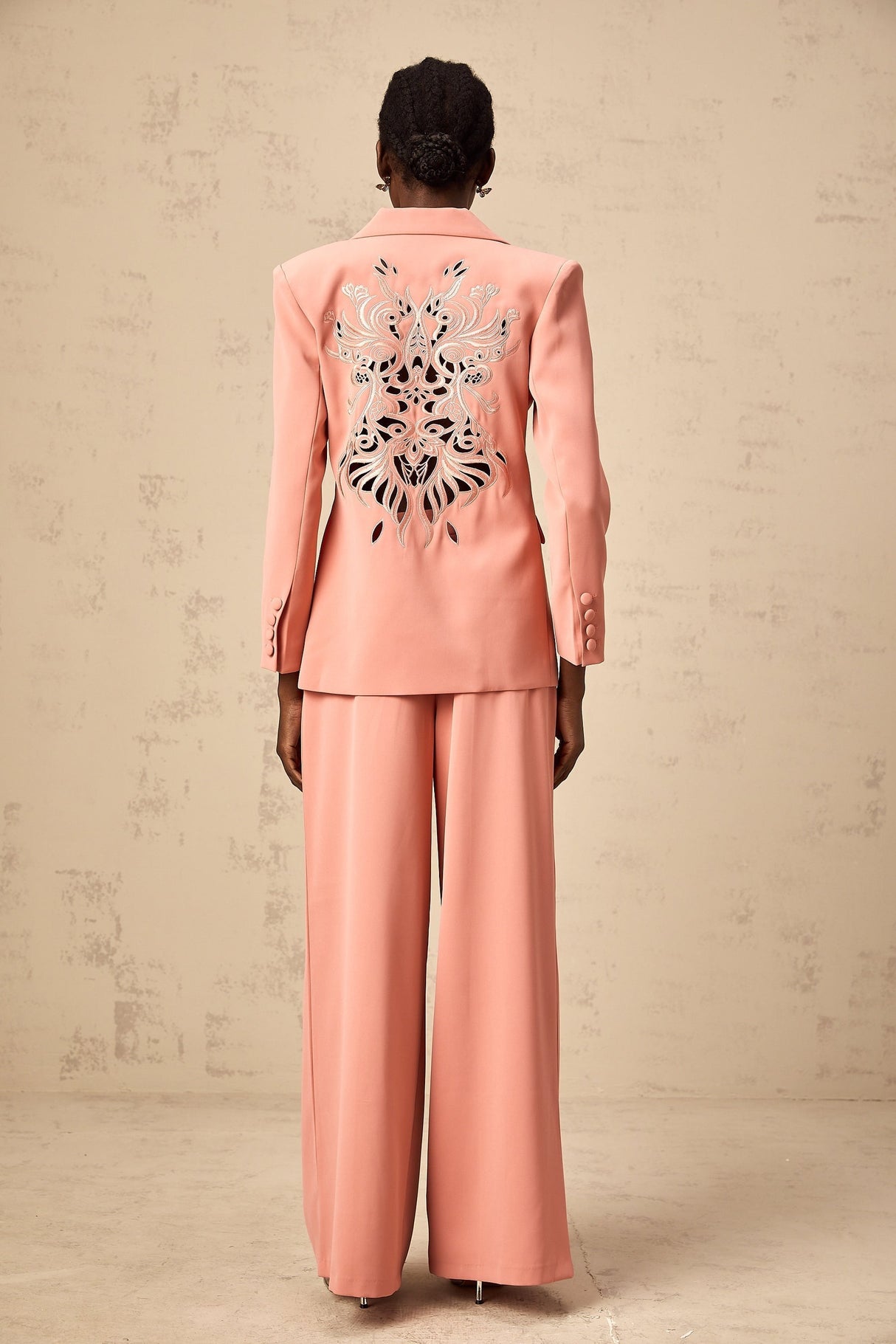 A pink blazer and trouser set with embroidery details and cutout designs The blazer features a tailored silhouette with decorative cutouts while the matching trousers have a sleek fit Both pieces are adorned with intricate pink embroidery creating an elegant and coordinated outfit