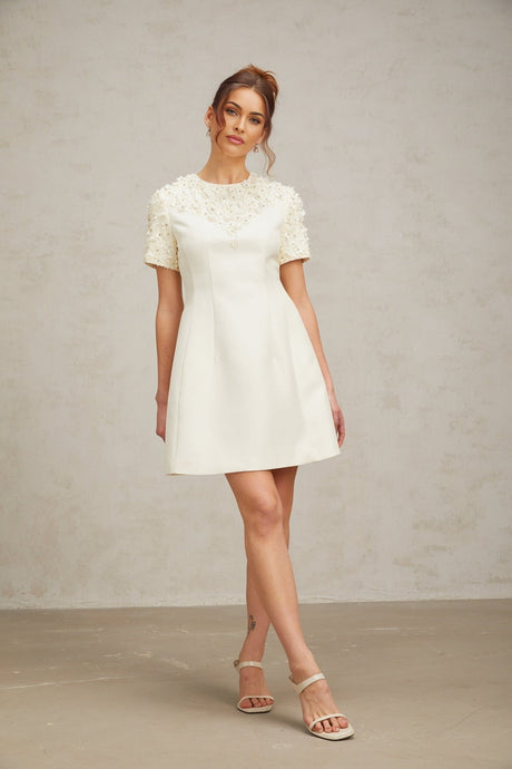 A short lightcolored mini dress with colorful floral appliqués decorating the fabric featuring a fitted bodice and flared skirt