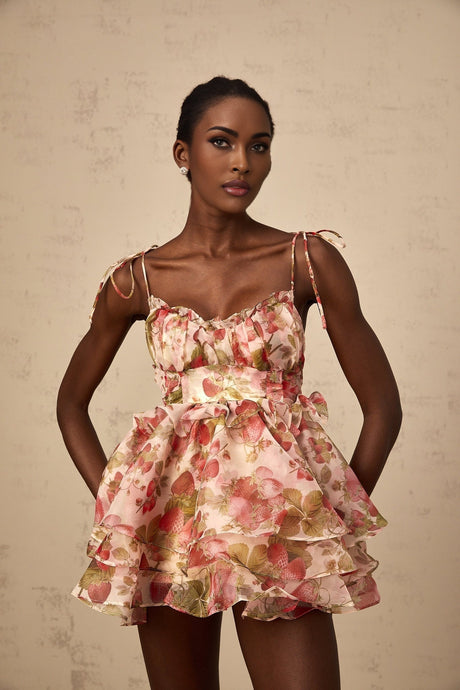 A short pink dress with a strawberry print pattern featuring a tiered skirt and likely short sleeves or straps styled in a feminine and playful design