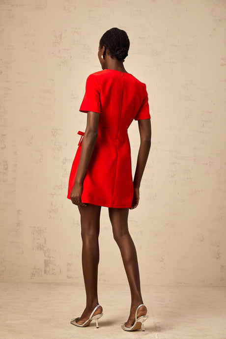 A short red mini dress with short sleeves and a bow detail worn by a model against a plain background