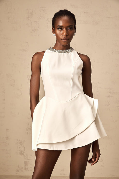 A short white mini dress with crystal embellishments and multiple layered tiers worn by a model against a plain background