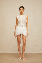 A short white mini dress with ruched fabric details and floral applique decorations on a model against a plain background