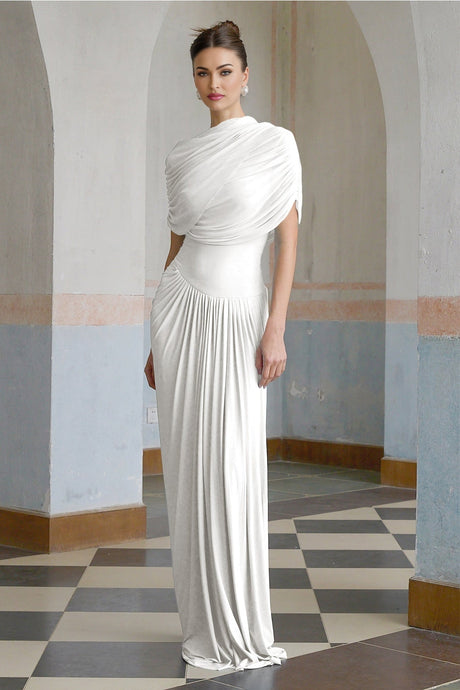 A white asymmetric pleated maxi dress with a long flowing skirt and uneven hemline modeled on a woman standing against a plain background