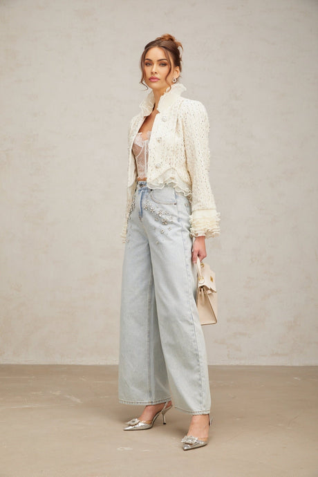 A white cropped jacket with beaded details and ruffle trim worn by a model against a plain background