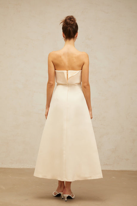 A white midi dress with a corsetstyle bodice and folded details worn by a model standing against a plain background
