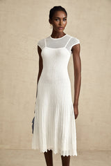A white midi dress with a scalloped hem and semi sheer fabric worn by a model standing against a plain background