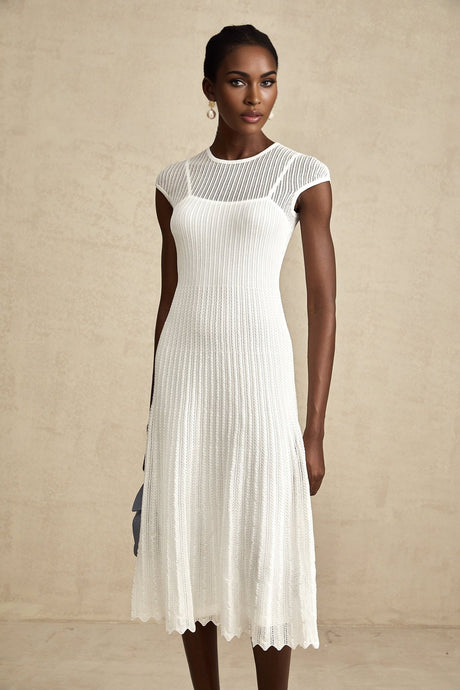 A white midi dress with a scalloped hem and semi sheer fabric worn by a model standing against a plain background