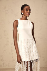 A white midi dress with discshaped embellishments and fringe details along the hemline worn by a model standing against a plain background