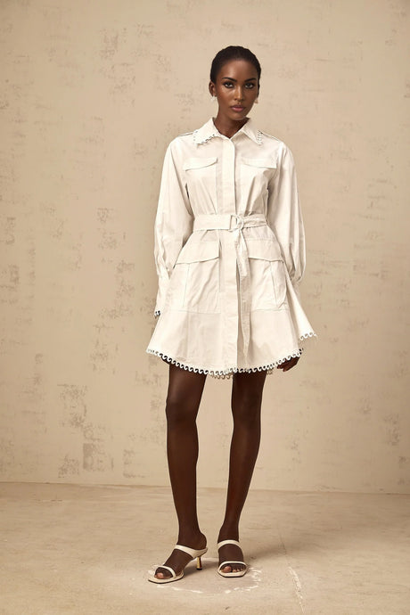 A white mini shirt dress with short sleeves and a belted waist worn by a model standing against a plain background