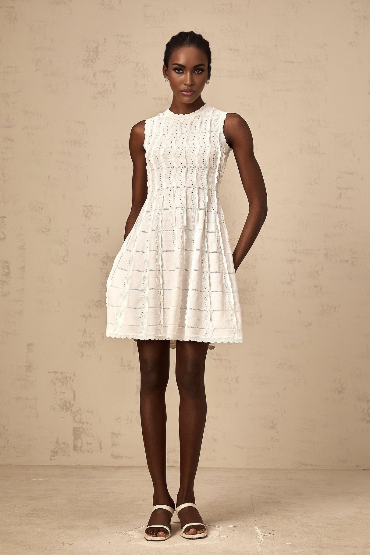 A white sleeveless mini dress with a knit fabric and ruffled details worn by a model standing against a plain background