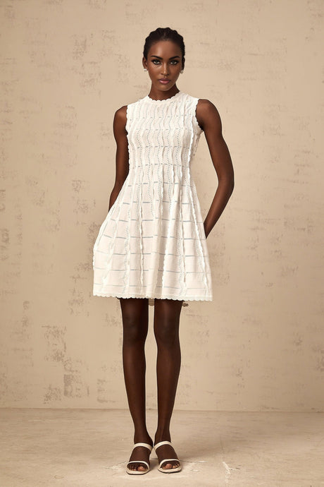 A white sleeveless mini dress with a knit fabric and ruffled details worn by a model standing against a plain background