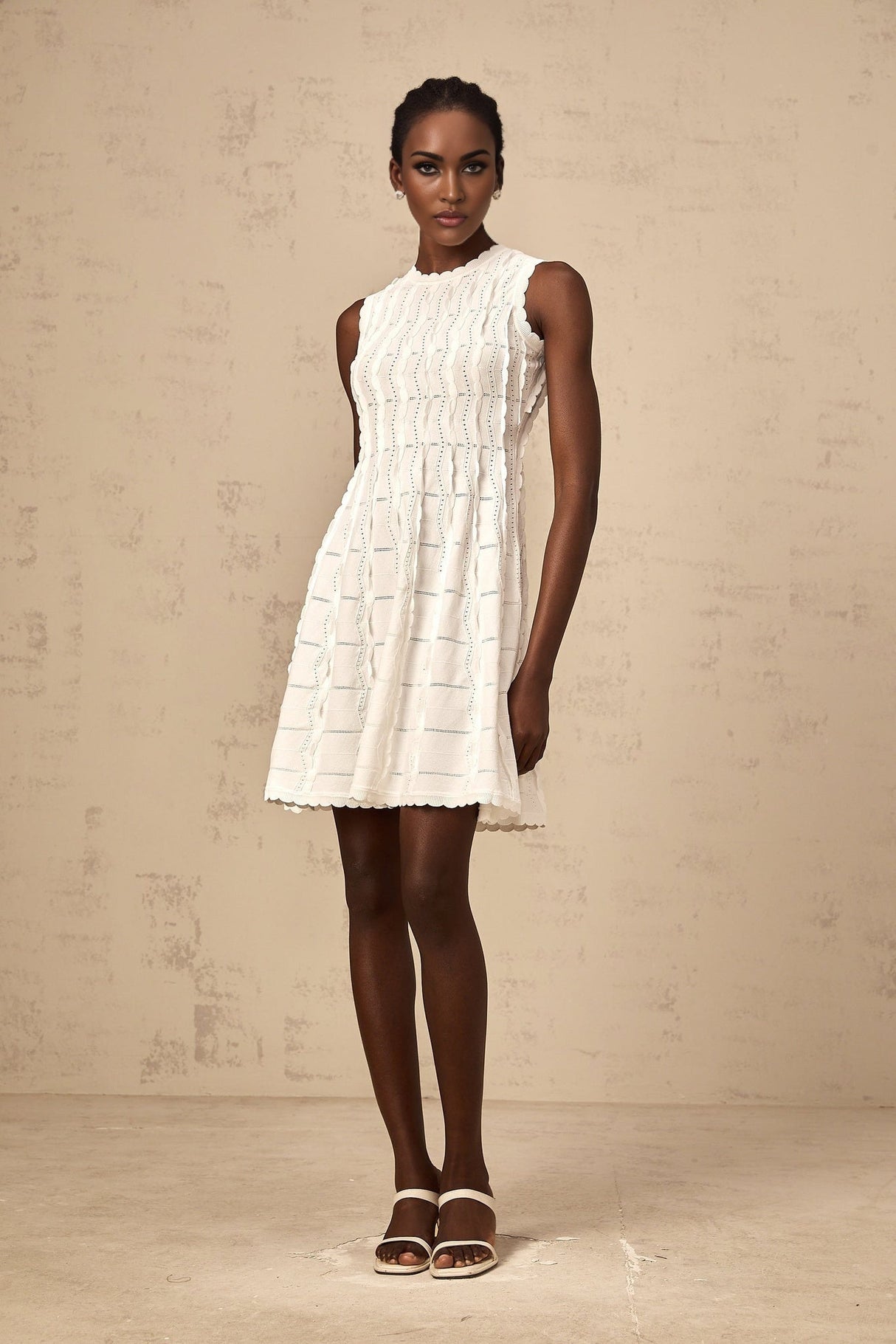 A white sleeveless mini dress with a knit texture and ruffled details worn by a model against a plain background
