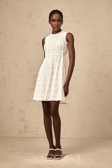 A white sleeveless mini dress with a knit texture and ruffled details worn by a model against a plain background