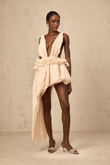 A woman wearing a beige mini dress with an asymmetric hemline and ribbon design details standing against a plain background