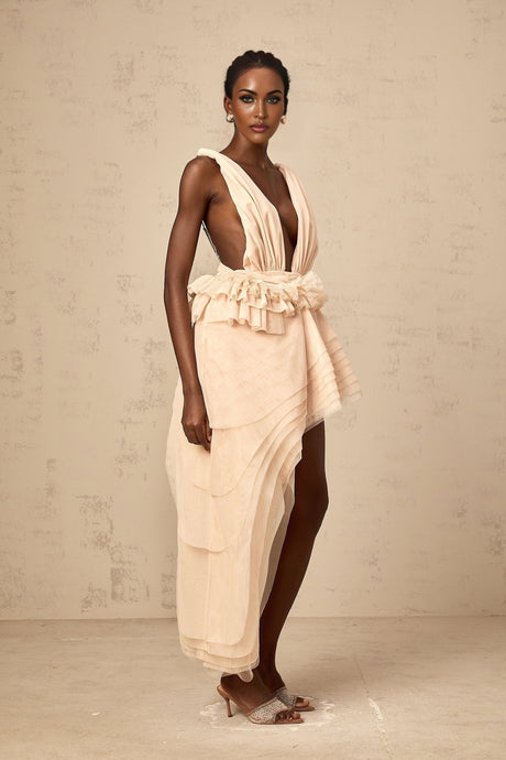 A woman wearing a beige mini dress with an asymmetric hemline and ribbon design details standing against a plain background