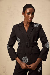 A woman wearing a black blazer and trousers set featuring studded rose details The blazer has a tailored fit and the trousers are slim cut The outfit is styled as a matching set