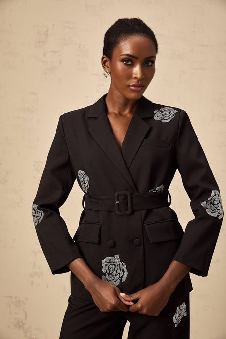 A woman wearing a black blazer and trousers set featuring studded rose details The blazer has a tailored fit and the trousers are slim cut The outfit is styled as a matching set
