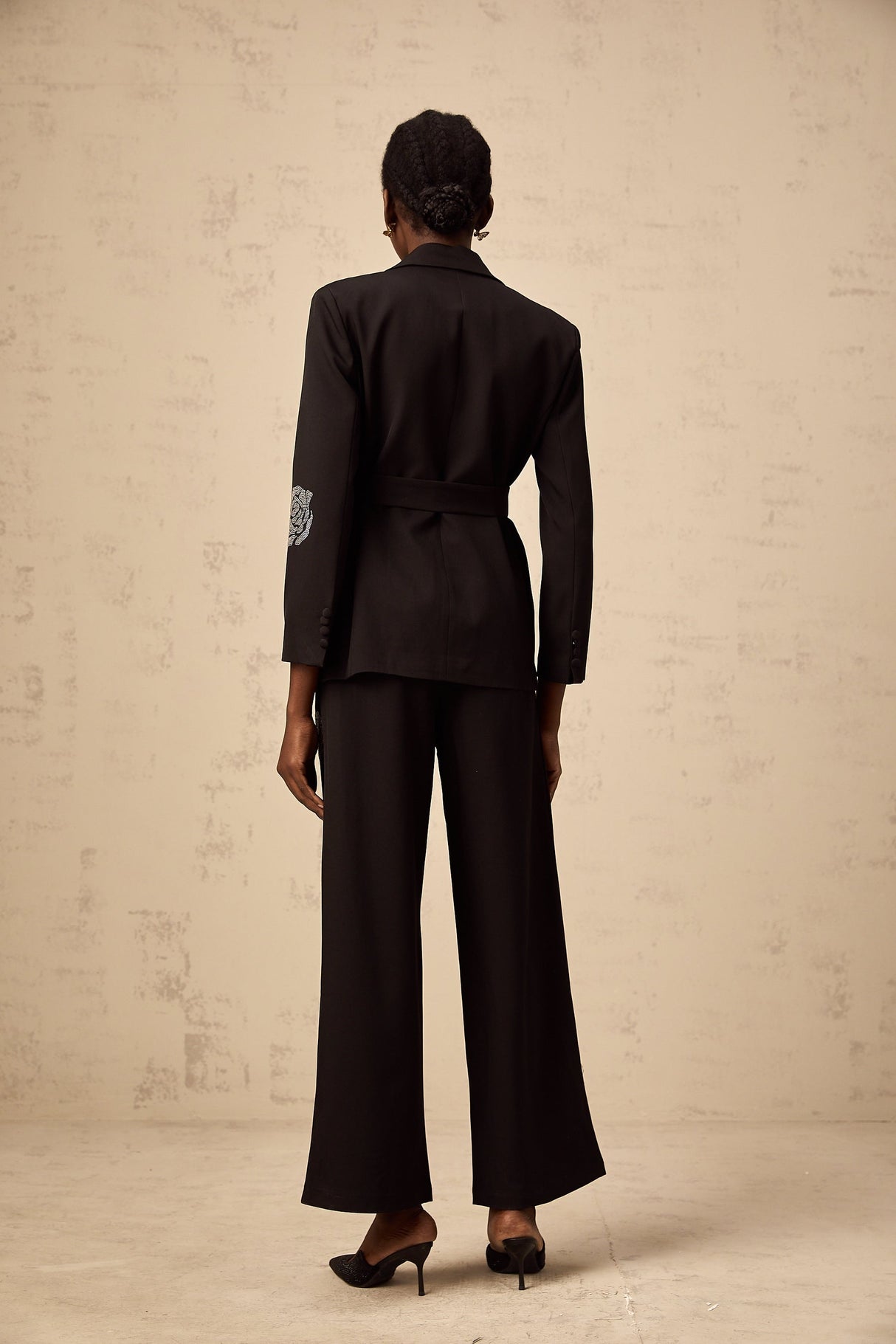 A woman wearing a black blazer and trousers set with studded rose designs The blazer has a tailored fit and the trousers are slim cut Both pieces feature decorative rose patterns made with small studs