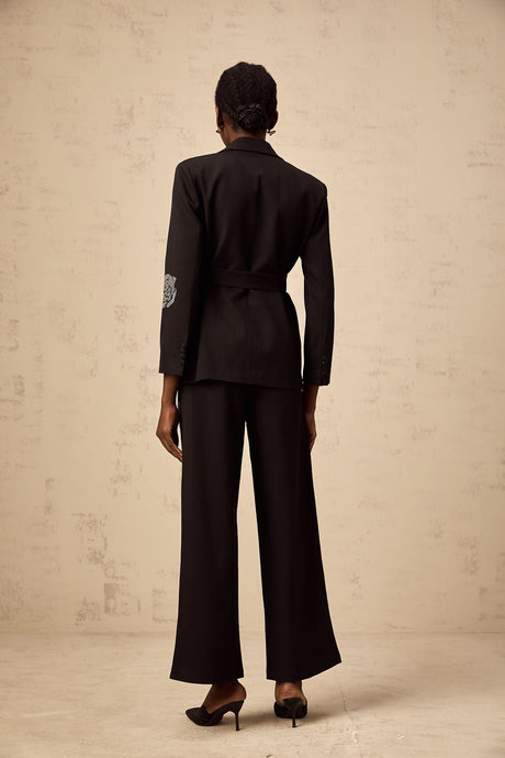 A woman wearing a black blazer and trousers set with studded rose designs The blazer has a tailored fit and the trousers are slim cut Both pieces feature decorative rose patterns made with small studs